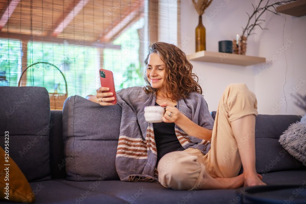 Smiling young woman relaxing on a couch, drinking coffee, using mobile phone