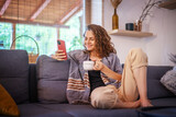 Smiling young woman relaxing on a couch, drinking coffee, using mobile phone