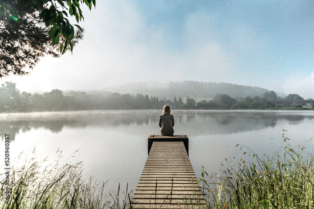 Calm misty morning relaxation by pond.Sitting woman in countryside.Intended female mysterious atmosphere. Spring foggy nature.Silence.Woman feeling freedom,enjoying vacation.No stress,calm mind,relax