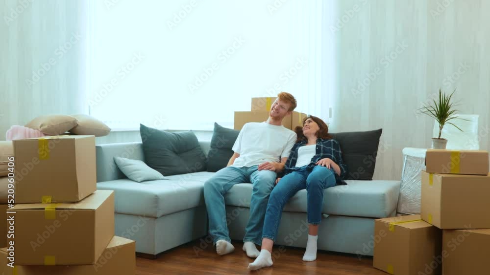 Caucasian joyful boyfriend and girlfriend moving in together in new house. Tired happy young married couple sitting down on sofa in room with many boxes on moving day. Home concept Caucasian joyful boyfriend and girlfriend moving in together in new house. Tired happy young married couple sitting down on sofa in room with many boxes on moving day. Home concept