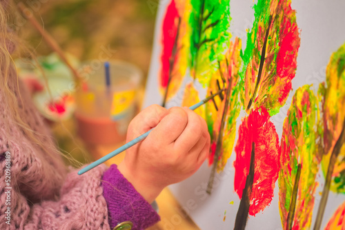 little girl artist with a brush and paints in her hands in autumn in the park draws a landscape with leaves on canvas.