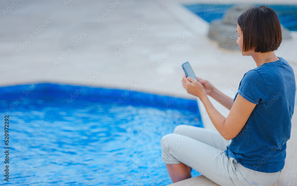 Young brunette woman using her phone while relaxing by the pool . Young lady downshifter working at phone enjoys and relaxed environment, working day. Online freelance work on vacation.