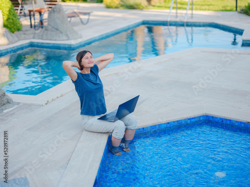 Young business woman working at computer by hotel pool. Young lady downshifter working at laptop and enjoys and relaxed environment, working day. Online freelance work on vacation.