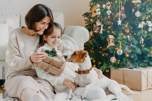 Happy young woman embraces her little daughter, holds Christmas present, awaits for winter holiday, play with pedigree puppy, pose on floor near decorated New Year tree. Happy family time concept