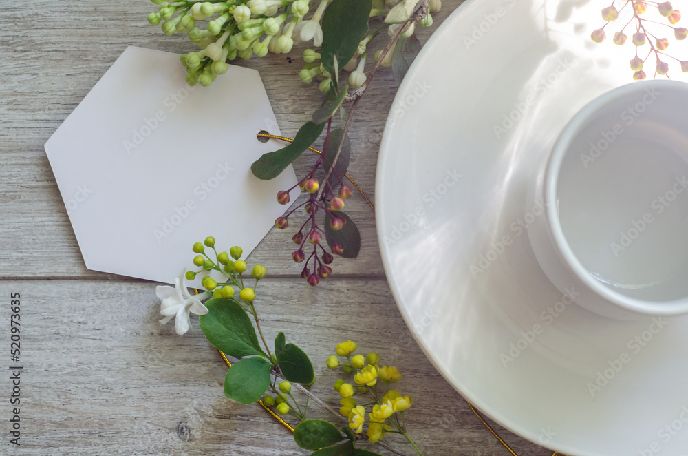 Flatlay close-up with an empty cup on a saucer with mockup an empty white card framed by branches of green leaves, white lilac flowers and yellow small flowers Flatlay close-up with an empty cup on a saucer with mockup an empty white card framed by branches of green leaves, white lilac flowers and yellow small flowers