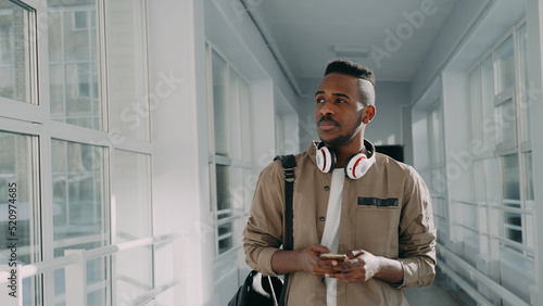 Young handsome african american male student with big white headphones walking in long lighty glassy corridor of college holding smartphone looking into it texting someone and smiling. photo