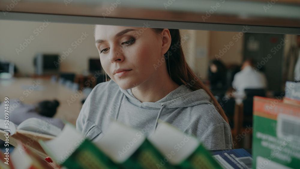 Reading Seriously Young caucasian beautiful female student is standing near shelf with ...