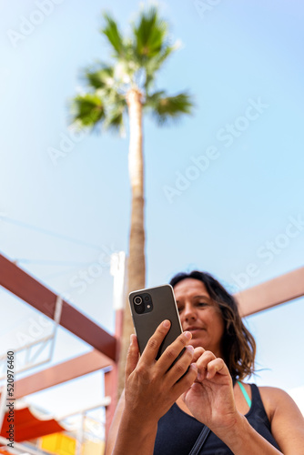 beautiful middle aged tourist using smartphone under a palm tree by the sea in exotic location