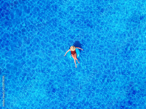 Zenith aerial view of a swimming pool in summer. Young girl in a swimsuit and hat floating with blue donut.