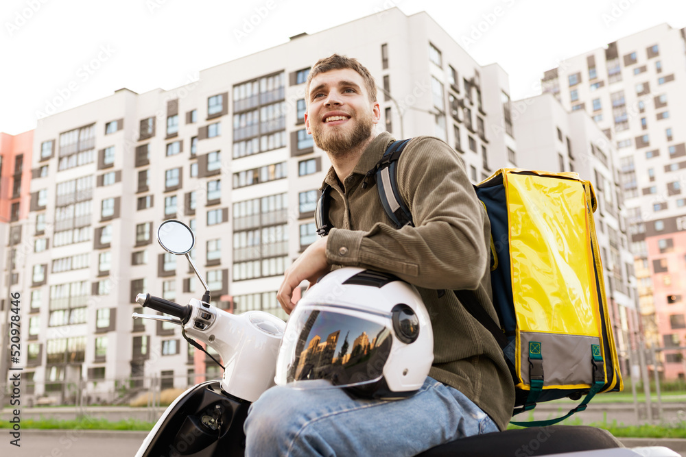 A smiling courier on a scooter with a yellow bag. The deliveryman parked near the house to deliver pizza to the customer - obrazy, fototapety, plakaty 