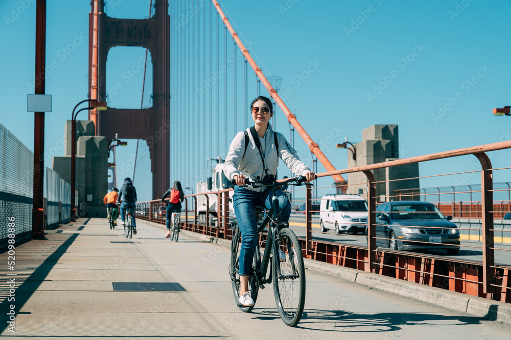 Fototapeta premium full length asian woman is enjoying cityscape while cycling at leisure on golden gate bridge in San Francisco California on a sunny day with vehicles passing by
