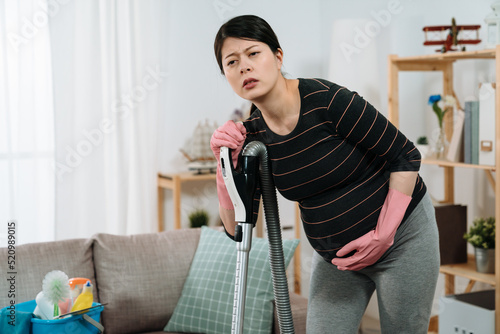 Photography portrait asian pregnant female bending and holding on a vacuum cleaner is suffering labor contraction pain with hand on the abdomen in the living room at home