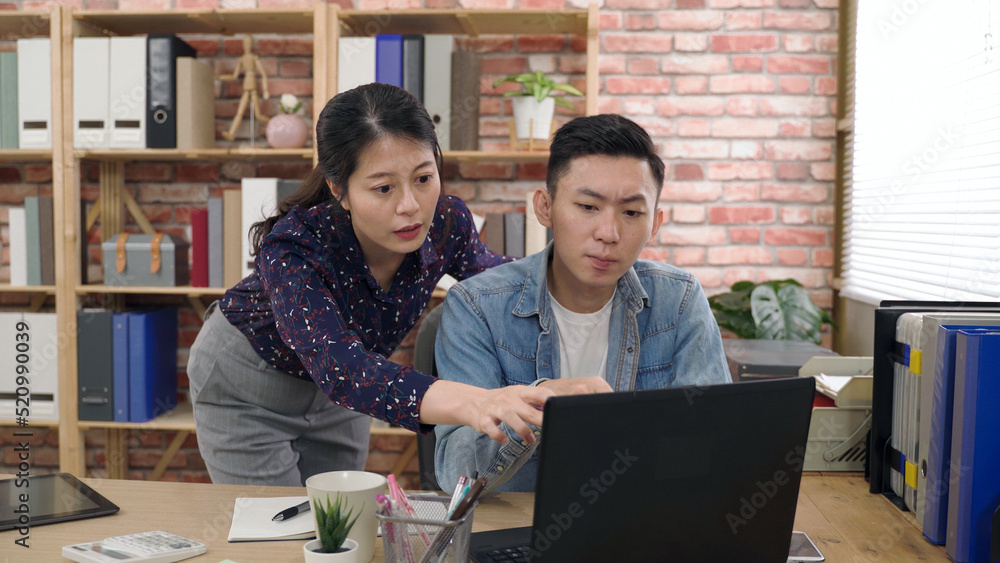 creative worker touching chin is nodding to show approval after his partner standing behind offers opinions by pointing at the monitor in the office. creative worker touching chin is nodding to show approval after his partner standing behind offers opinions by pointing at the monitor in the office.