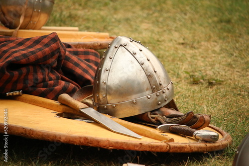 Equipment, costume and props of a Viking reenator lying on the ground outdoors; helmet, wooden handled knife, sword, shield and tunic.