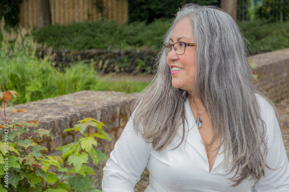 Naklejka premium Profile portrait of a mature woman with a calm smiling expression, tanned skin, straight gray hair, glasses, light makeup, sunny day in the garden with green vegetation in the background