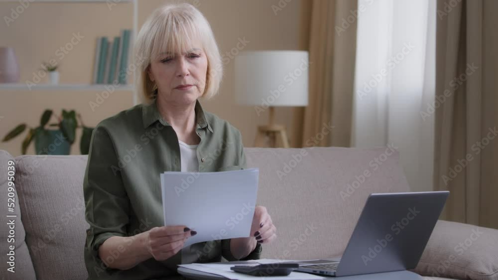Caucasian old 50s homeowner housewife makes woman budget checks monthly income by checking paper notes using app laptop. Middle-aged businesswoman sitting on couch with document working remote at home Caucasian old 50s homeowner housewife makes woman budget checks monthly income by checking paper notes using app laptop. Middle-aged businesswoman sitting on couch with document working remote at home