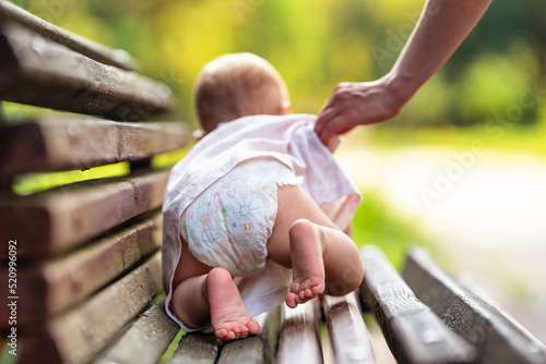Baby in diaper crawling on a park bench on a sunny summer day. Mother's hand helps and protects the child. Happy kid learn and play.
