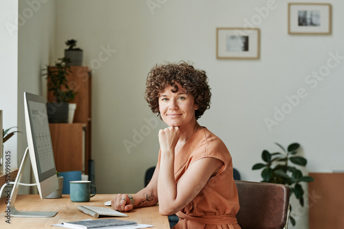 Portrait of young businesswoman sitting at her workplace in front of computer monitor at office and looking at camera