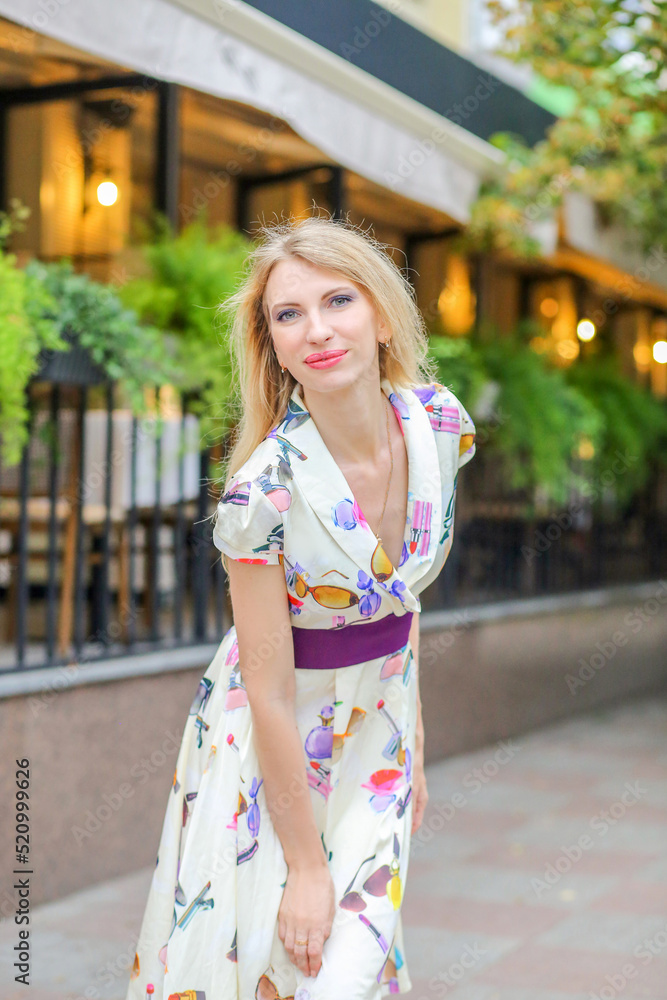 Portrait of a happy young blonde girl smiling at the camera on the background of a summer city street. Beautiful summer dress. Beauty and fashion concept