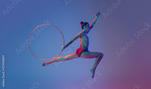 Portrait of young sportive girl, female rhythmic gymnast training, performing with hoop isolated on blue purple background in neon light © Lustre