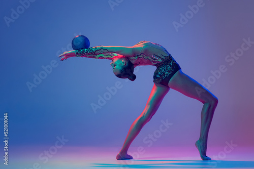 Portrait of young girl, rhythmic gymnast athlete performing with blue ball isolated on blue purple background in neon light © Lustre Art Group 