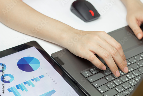 Closeup of female hand holding a pen and typing on a computer keyboard while looking at graphs.