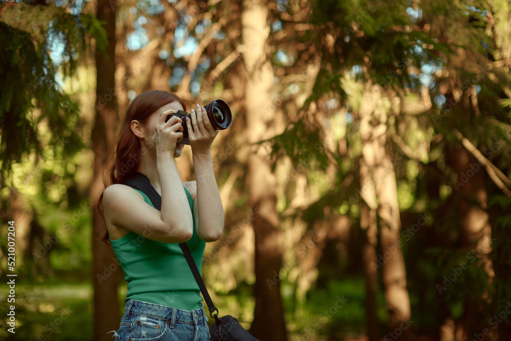 A young red-haired woman in a green top and denim shorts takes pictures in the woods.