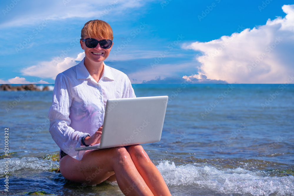 Woman working remotely and using a laptop during summer vacation on sea. Copy space.