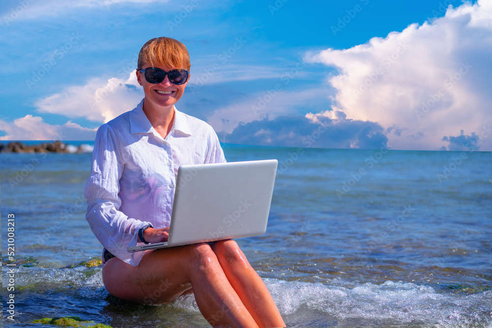 Woman working remotely and using a laptop during summer vacation on sea. Holiday, business,  quality of life concept. Horizontal image.