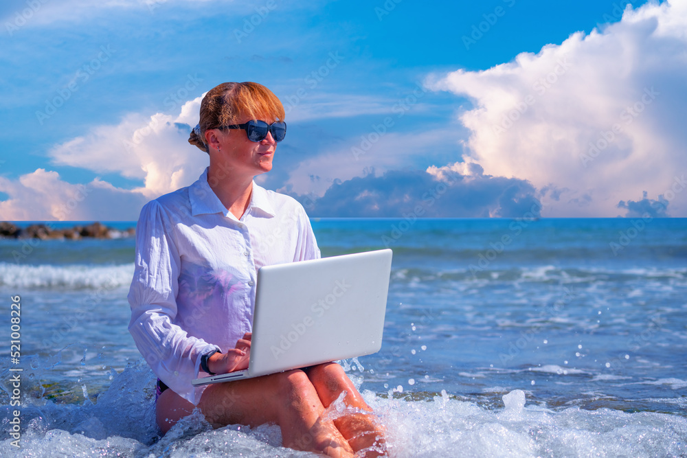 Beautiful woman working remotely and using a laptop during summer vacation on sea. Holiday, business,  quality of life concept.