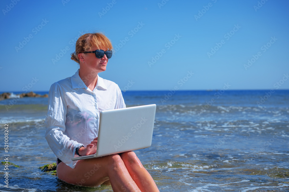 Woman working remotely and using a laptop during summer vacation on sea. Holiday, business,  quality of life concept. Copy space.