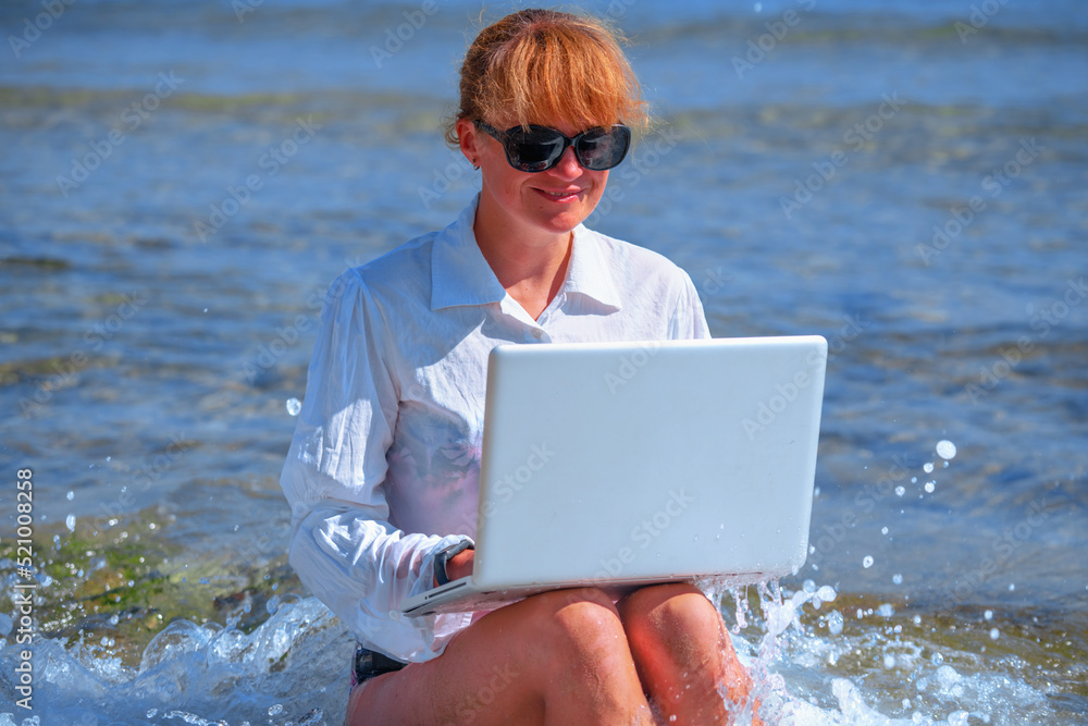 Woman working remotely and using a laptop during summer vacation on sea. Holiday, business,  quality of life concept.