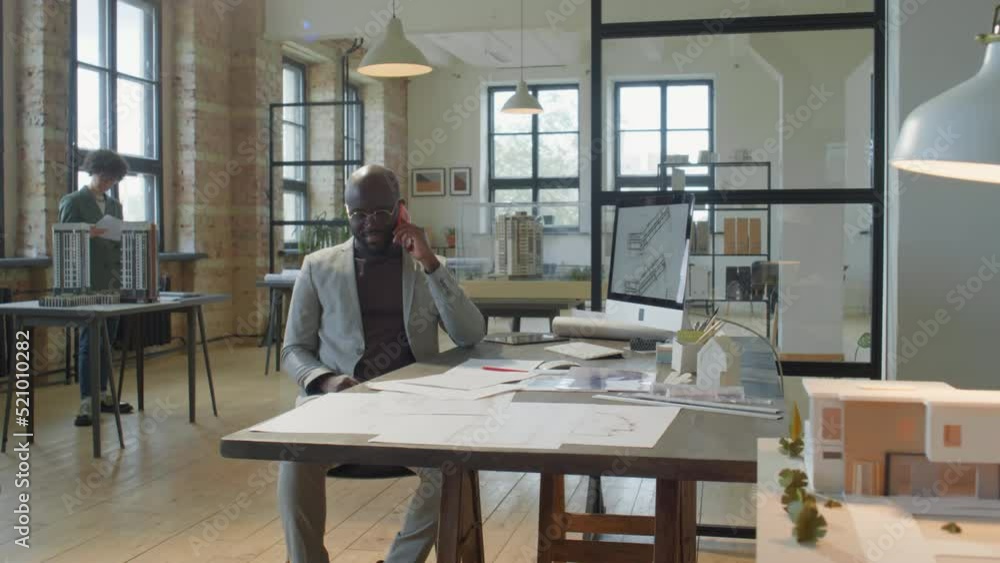 Zoom in shot of African American businessman sitting at office desk and speaking on mobile phone during workday in architectural office Zoom in shot of African American businessman sitting at office desk and speaking on mobile phone during workday in architectural office