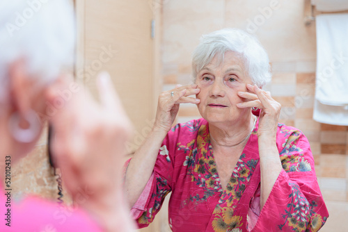 Mature woman taking care of her face. Senior woman standing at the mirror in the bathroom and applying cream to her face or doing self-massage