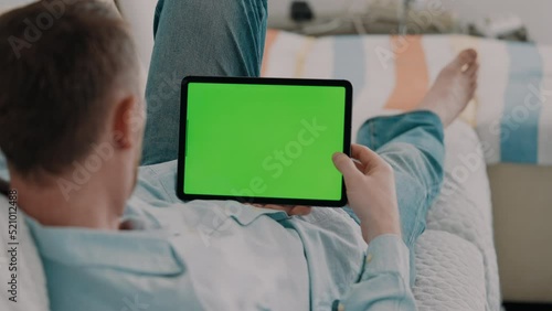 leisure time at home man lying on a couch wearing casual shirt holding digital tablet horizontally scrolling on green screen