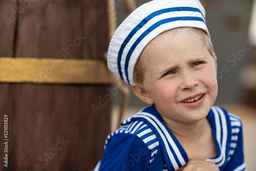 A close-up emotional portrait of a boy in a sailor suit and cap. The boy is sitting on the background of a brown wooden barrel. Summer, wood flooring. Emotions of a child