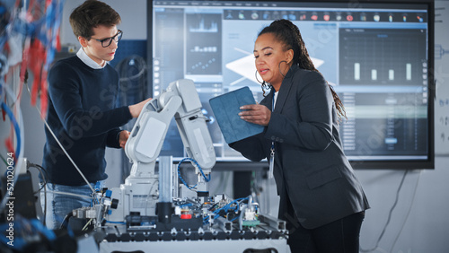 Black Female Chief Engineer Holding Tablet and Talking with Male Student while Robot Arm Moving Under their Supervision During the Lesson. Computer Science Education and Learning Concept.