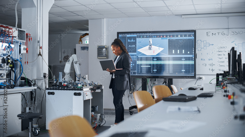 Fototapeta premium Black Female Automation Engineer Holding Laptop and Moving Bionic Arm With Written Code at Futuristic Factory. Modern Equipment and New Era in Computer Science and Robotics Industry Concept.