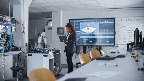 Black Female Automation Engineer Holding Laptop and Moving Bionic Arm With Written Code at Futuristic Factory. Modern Equipment and New Era in Computer Science and Robotics Industry Concept.