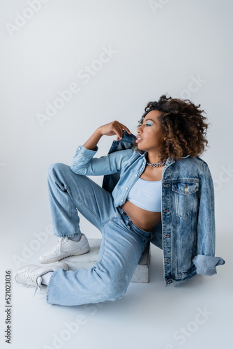 full length of stylish african american woman in white sneakers and blue denim clothes sitting on grey background © LIGHTFIELD STUDIOS