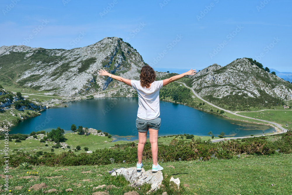 40-year-old brown-haired girl from the back with open arms looking towards a mountain lake in the background