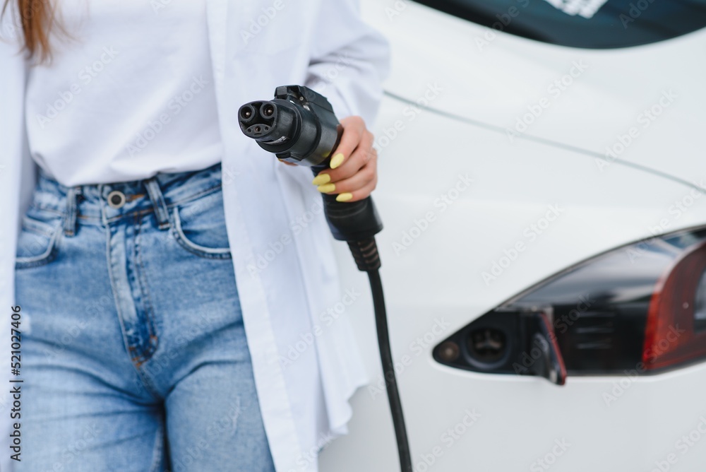 A girl's hand holds a cable for charging an electric car on the background of a white car. A girl's hand holds a cable for charging an electric car on the background of a white car.