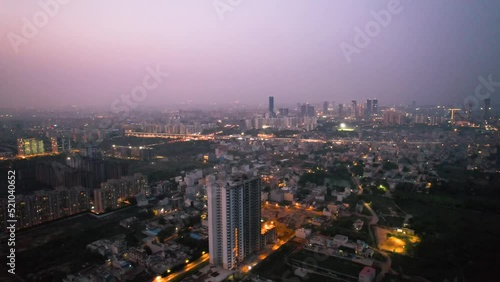 aerial drone dusk twilight shot showing orange lights of streets, homes and markets surrounding a skyscraper with the city scape stretching into the distance in gurgaon haryana delhi