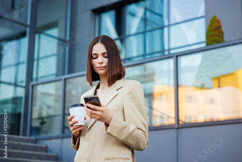 A young brunette girl against the backdrop of a business center, office center. Portrait of a successful startup. Smartphone, coffee casual suit.