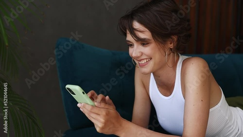 Young dreamful pensive minded woman wears casual clothes lying on blue sofa hold using mobile cell phone typing chat send sms stay at home rest relax spend free time in living room indoors grey wall