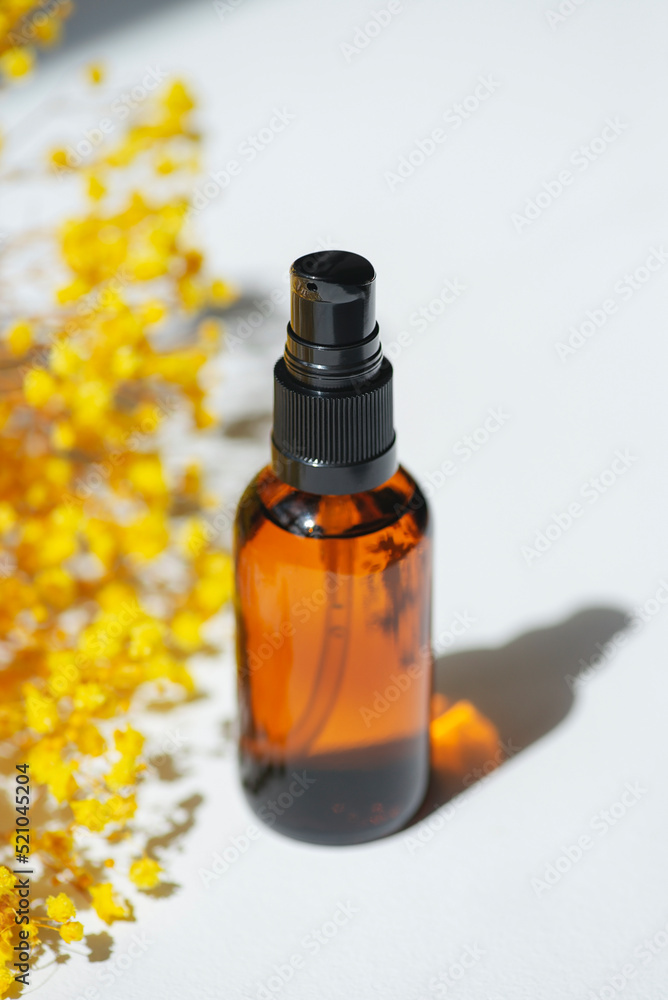 Amber pump bottle with serum, tonic or essential oil with yellow flowers. White background with daylight. Beauty concept for face and body care Amber pump bottle with serum, tonic or essential oil with yellow flowers. White background with daylight. Beauty concept for face and body care