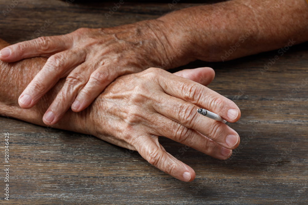 Elderly wrinkled woman hands with a cigarette on wooden table. Concepts of women's health, bad habits, lung cancer disease. Shallow depth of field Elderly wrinkled woman hands with a cigarette on wooden table. Concepts of women's health, bad habits, lung cancer disease. Shallow depth of field