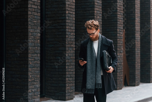 Holding smartphone. Stylish man with beard and in glasses is outdoors near building