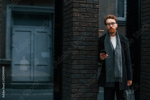 Holding smartphone. Stylish man with beard and in glasses is outdoors near building © standret