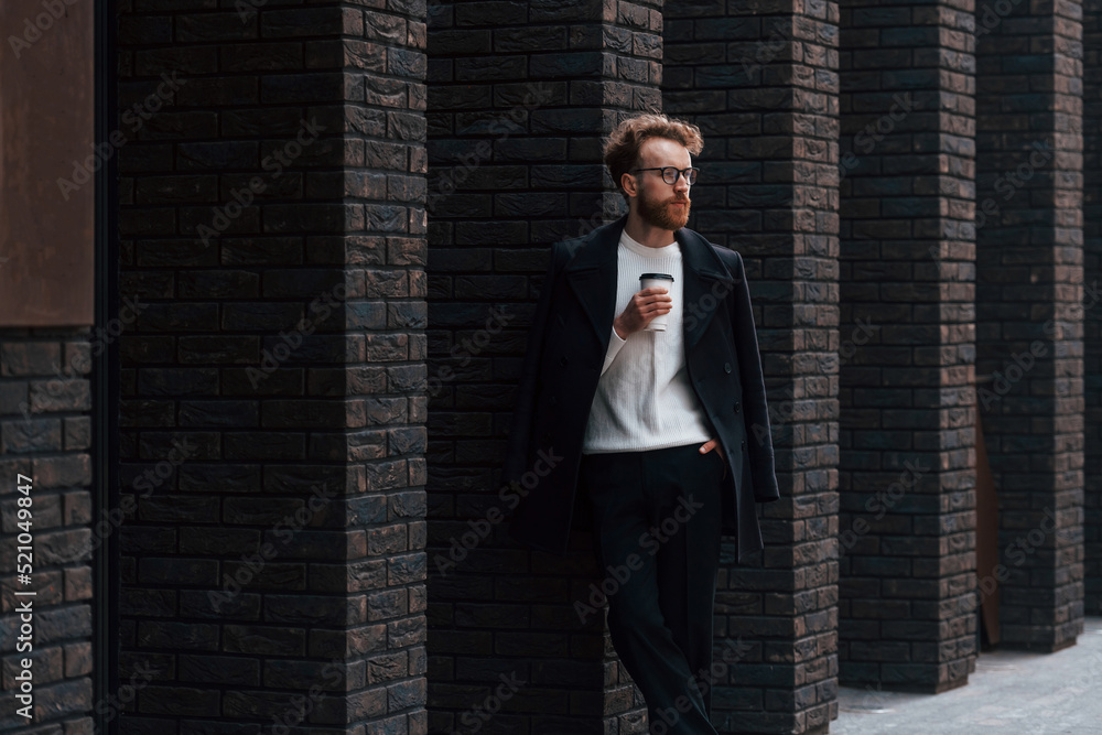 Standing with cup of coffee. Stylish man with beard and in glasses is outdoors near building Standing with cup of coffee. Stylish man with beard and in glasses is outdoors near building